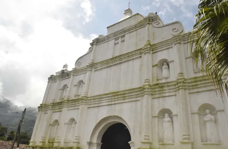 The white colonial facade of a village church with saints in niches and a palm leaning in from the right.