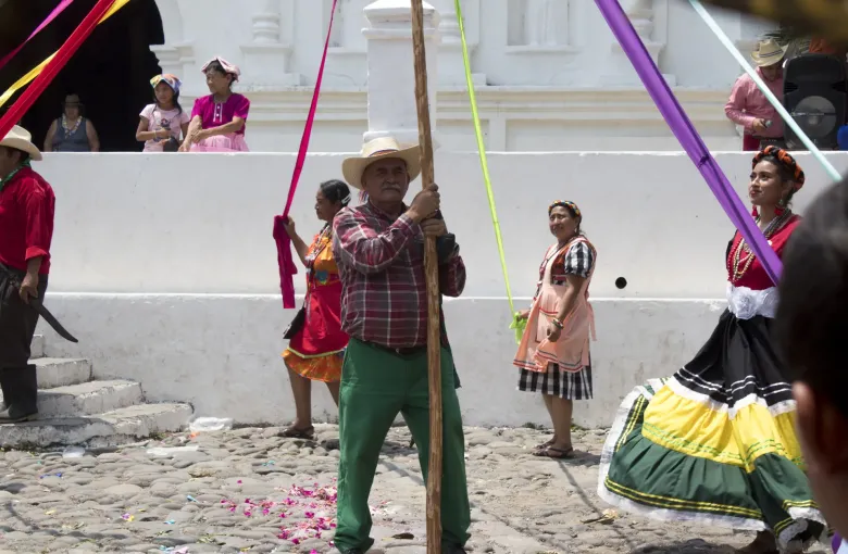 Dancers in bright layered skirts circle a ribboned pole held upright by a man in a straw hat on a cobbled plaza.