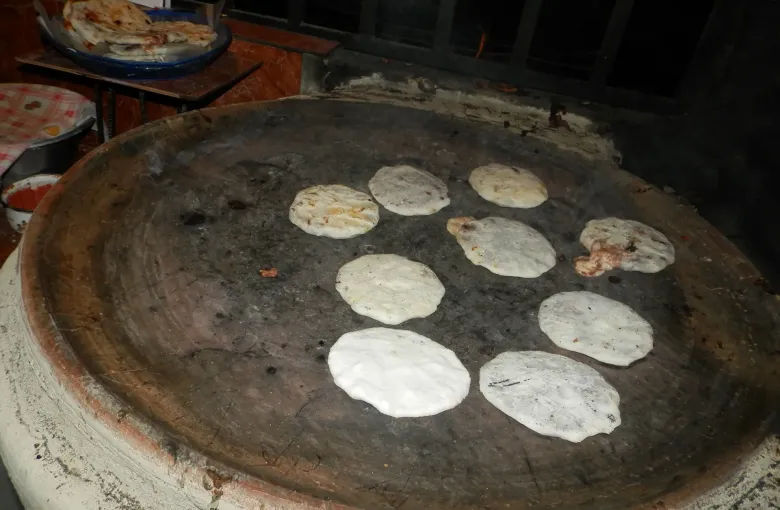 Eight pupusas cooking on a large round clay griddle over a fire, each one browning in patches.
