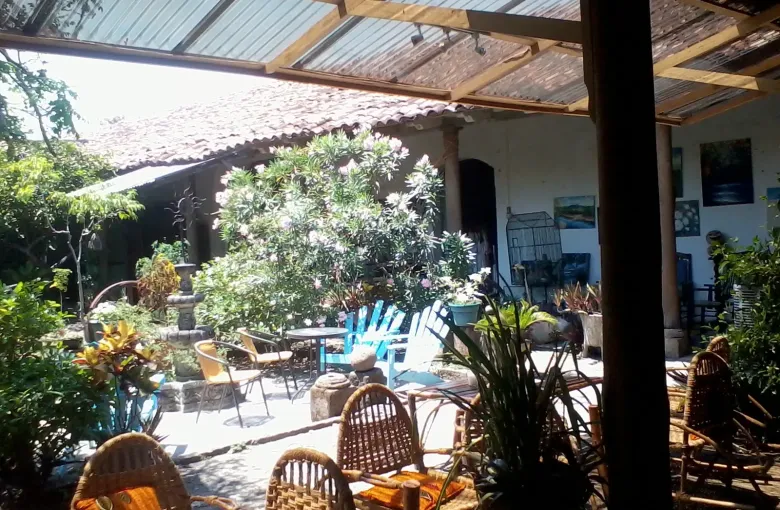 Wicker and wooden chairs set out under a corrugated canopy beside a planted courtyard, with a tiled roof behind.