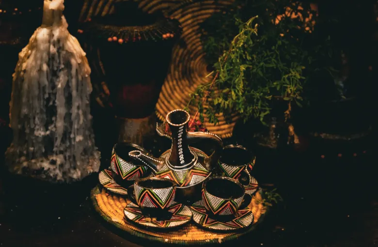 A clay jebena and a set of small handleless cups arranged on a decorated tray for a coffee ceremony, with grass strewn around them.