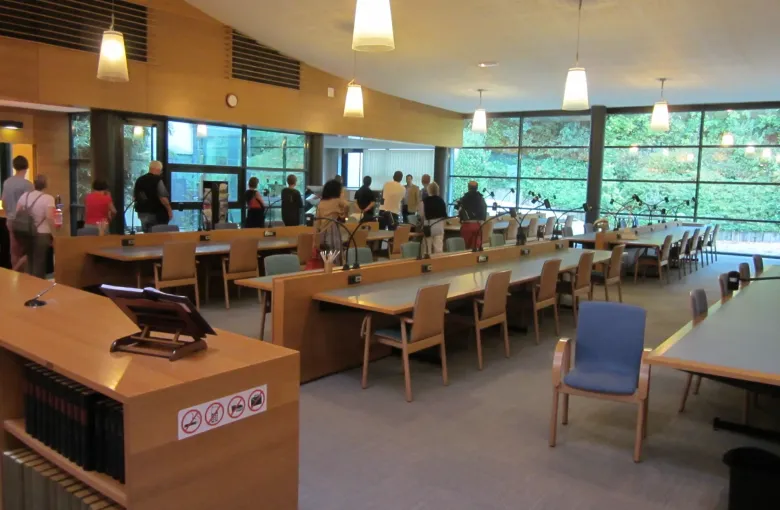 Long reading tables with individual lamps and bound registers on a shelf in a French departmental archives room.