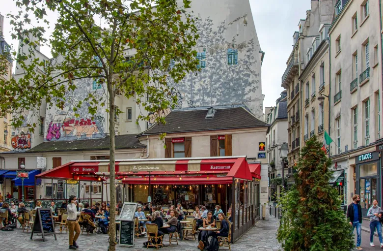 A full cafe terrace under red awnings on a small Paris square, with tables close together and the street beyond.