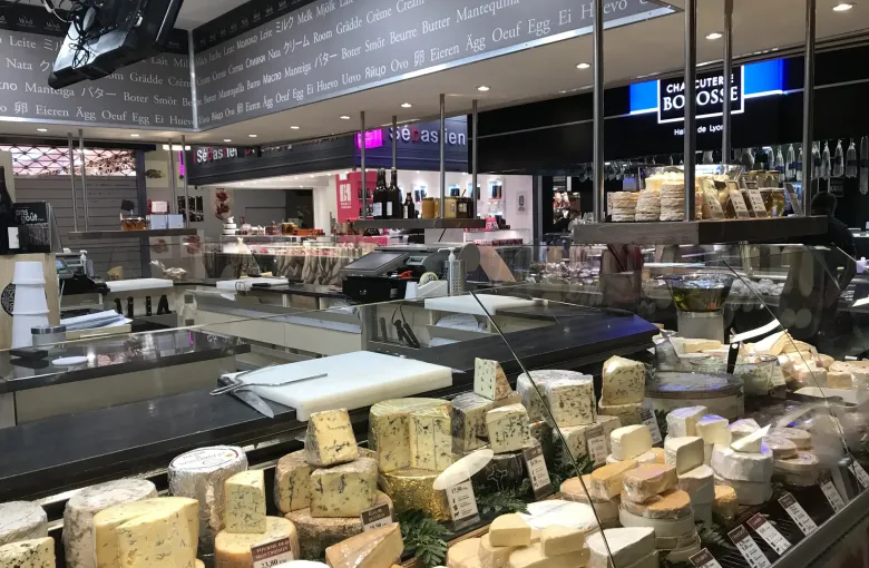 Wheels and rounds of cheese massed across a covered market counter under a hand lettered board in Lyon, France.
