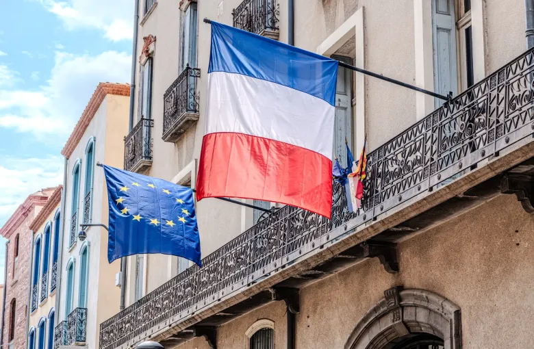 The French tricolour and the European Union flag hanging side by side from an ironwork balcony on a French street.