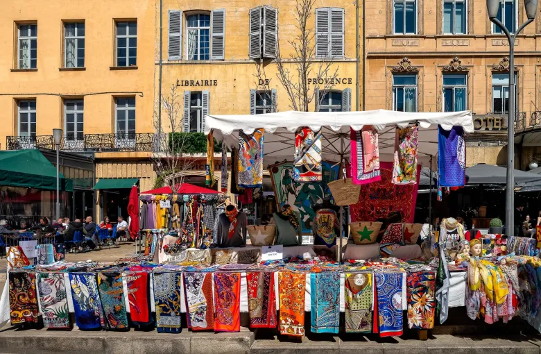 A market stall hung with printed scarves and textiles under a white awning on a shaded square in Provence, France.