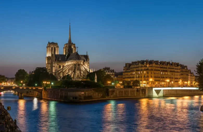 The spire and flank of Notre Dame above the lit quays of the Ile de la Cite, reflected in the Seine at dusk in Paris.