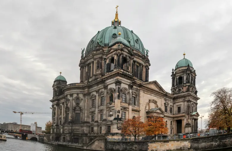 The green dome of Berlin Cathedral above the Spree embankment, with autumn trees along the water in front of it.