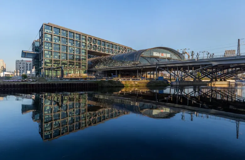 The glass hall of Berlin Hauptbahnhof reflected in the still water of the Spree, with the elevated tracks running through it.