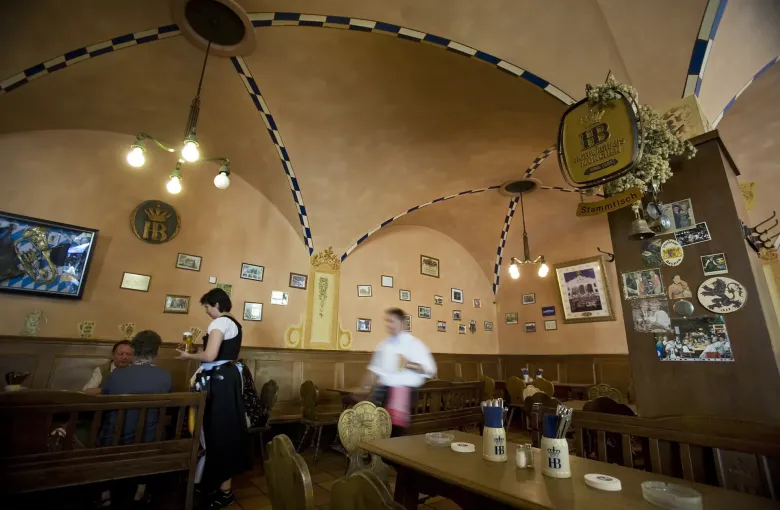 The vaulted interior of a Munich beer hall with long shared tables, a server carrying steins and the walls hung with prints.