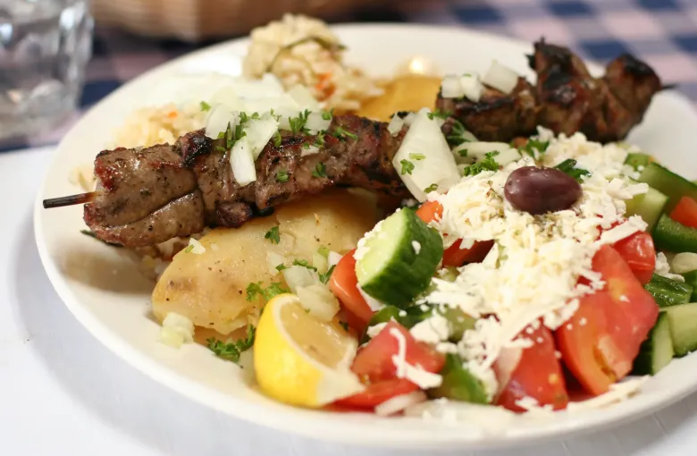 A plate of lamb souvlaki with potatoes, tzatziki and a Greek salad of tomato, cucumber and grated cheese.