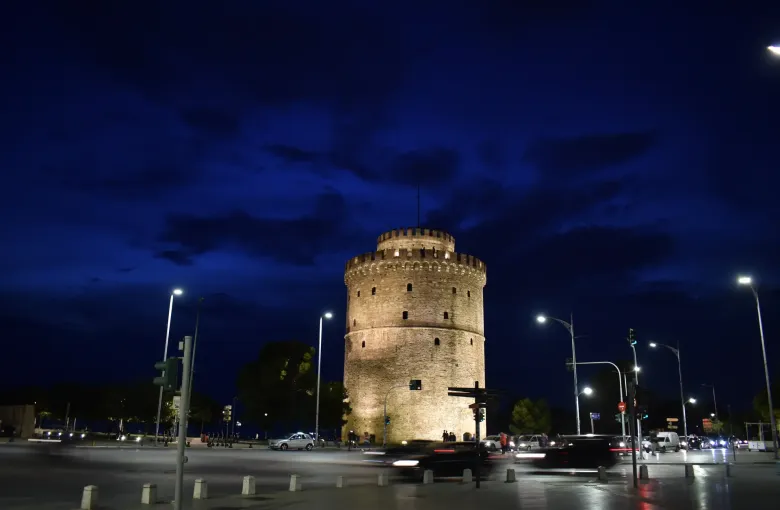 The floodlit White Tower of Thessaloniki against a deep blue evening sky, with traffic passing in front.