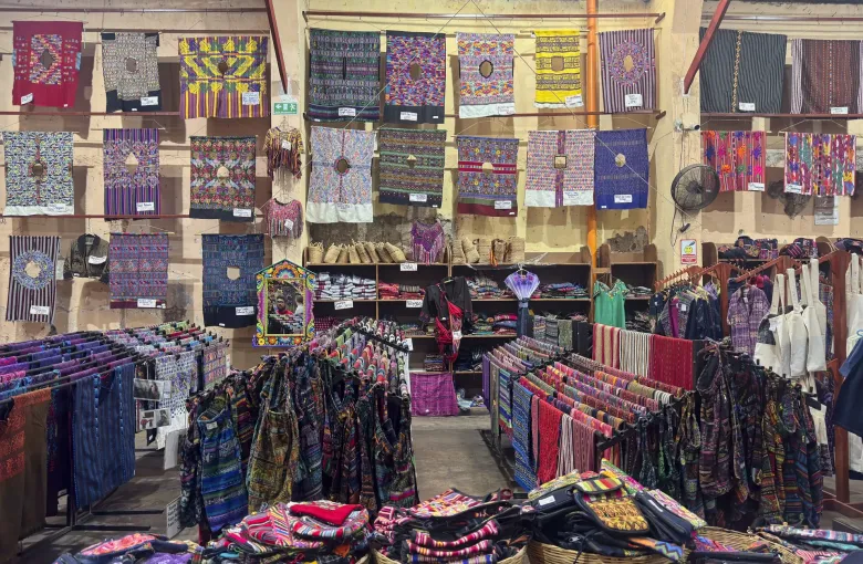 The interior of an artisan market in Antigua, its walls hung with woven huipiles and its floor filled with baskets of textiles.