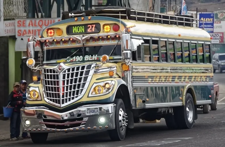 A repainted former school bus, chrome-trimmed and lit, working a street route in Guatemala with its destination board lit above the windscreen.