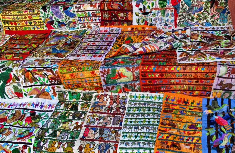 Layers of brightly embroidered Guatemalan cloth worked with birds, animals and human figures, stacked for sale.