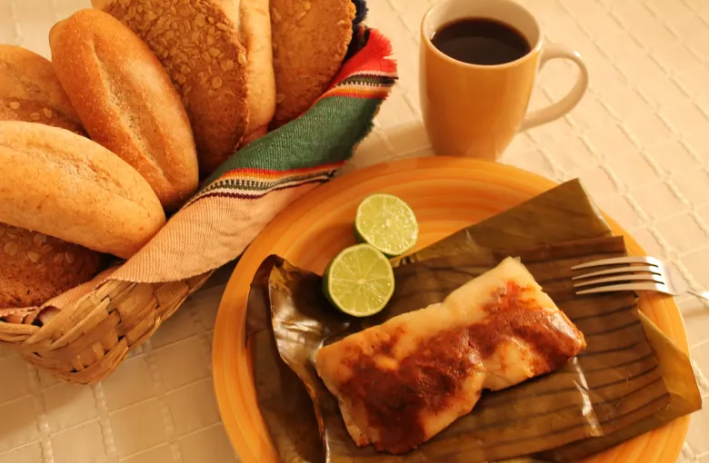 A Guatemalan tamal opened on its banana leaf beside halved limes, a basket of bread rolls and a cup of black coffee.