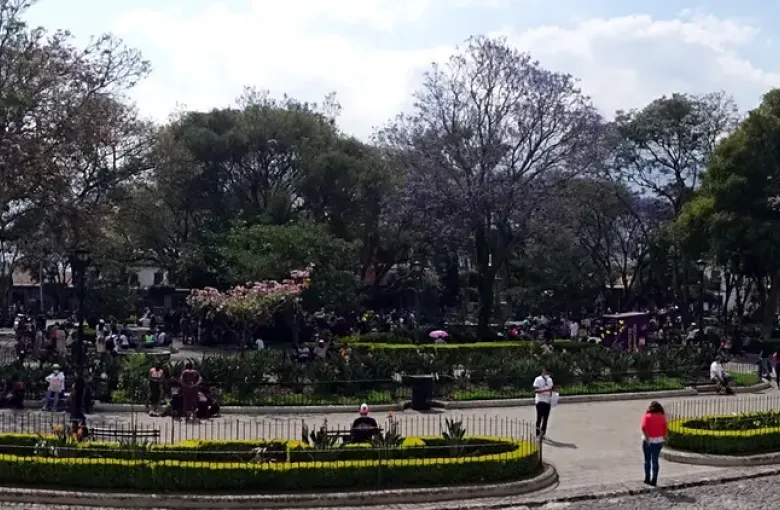 A wide view across Plaza Mayor in Antigua, showing flat paved walkways between the central gardens and the colonnaded buildings around it.