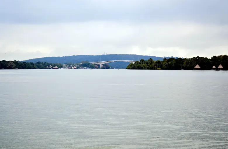 The long road bridge over the Rio Dulce seen from the water, with thatched shelters along the far bank under grey cloud.