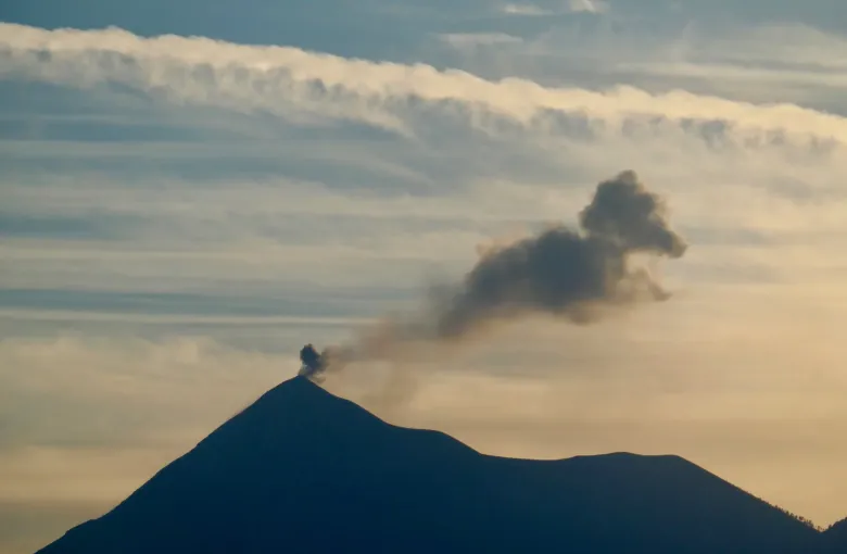 The silhouette of Volcan de Fuego at dusk with an ash plume drifting from its summit across a pale sky.