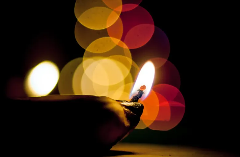 A lit clay diya burning against a dark background with warm circles of light behind it.
