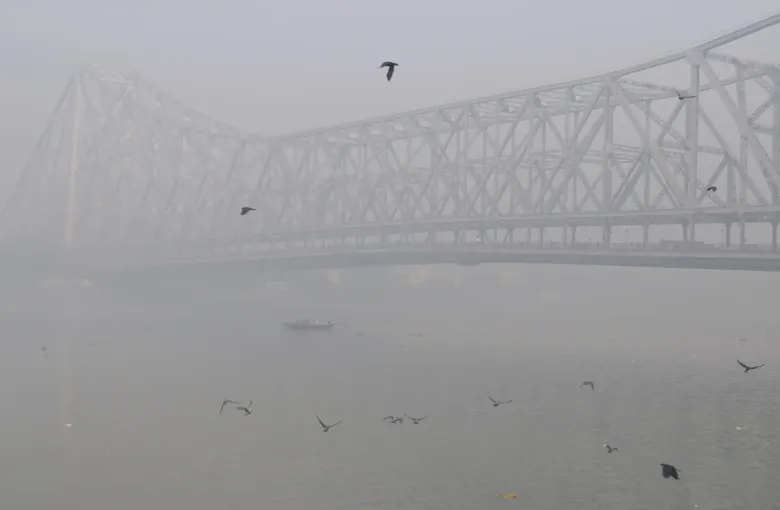 Howrah Bridge in Kolkata seen through winter fog above the Hooghly, with birds over the water.