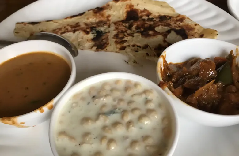 Flatbread, dal, a yogurt raita and a dark vegetable curry served together in white bowls.