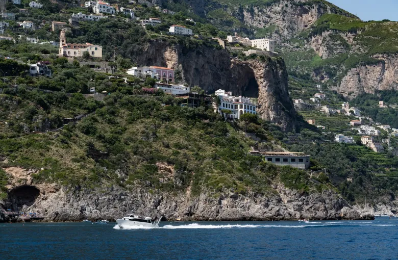 The cliffs and pastel houses of the Amalfi Coast seen from the water, with a boat passing below.