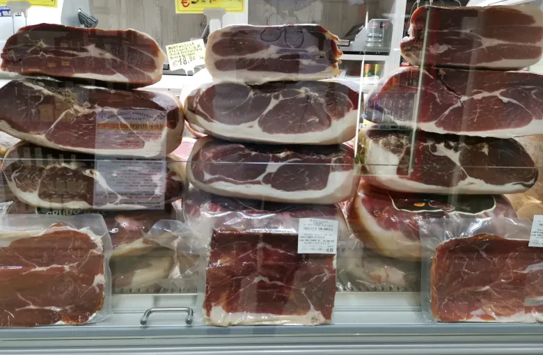 Whole cured hams stacked in a market case behind handwritten price tags in euros, Florence.