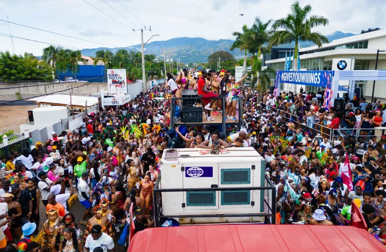 A music truck leads a dense carnival crowd along a Jamaican street, with hills on the skyline.