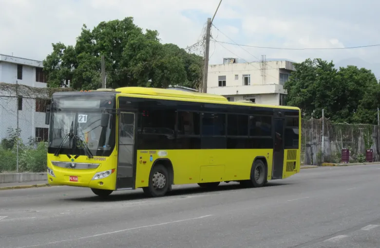 A yellow Jamaica Urban Transit Company city bus on route 21 runs along a Kingston street.