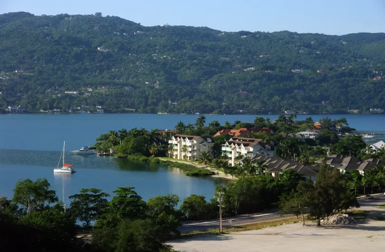 A catamaran lies at anchor off a green headland of houses on the Montego Bay shoreline.