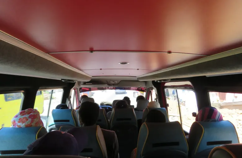 The interior of a taxi-brousse minibus seen from the rear seat, passengers facing forward as the vehicle waits at a roadside stop.
