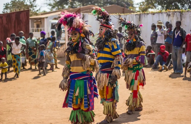 Masked Gule Wamkulu dancers in tall feathered headdresses and fringed costumes performing on open ground, with a crowd watching behind.