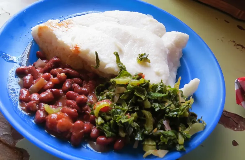 A blue enamel plate holding two portions of white nsima with stewed beans, cooked greens and a piece of fish.