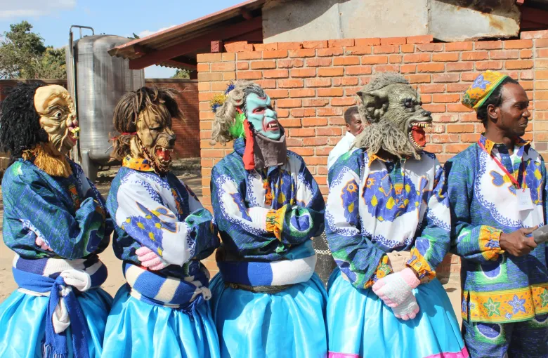 A line of Nyau dancers in blue and white satin costumes and carved masks standing in front of a brick building.