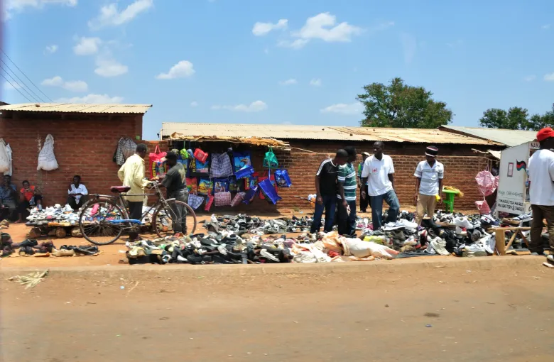A roadside stall in Malawi with clothing and shoes laid out on the ground in front of a brick building, and customers standing along the roadside.