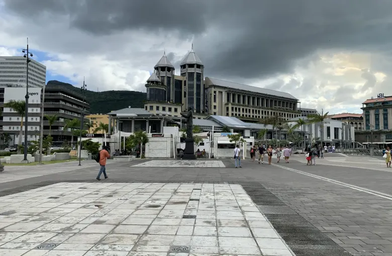 The broad level paving of the Caudan Waterfront plaza in Port Louis under heavy cloud, with people crossing the open square.