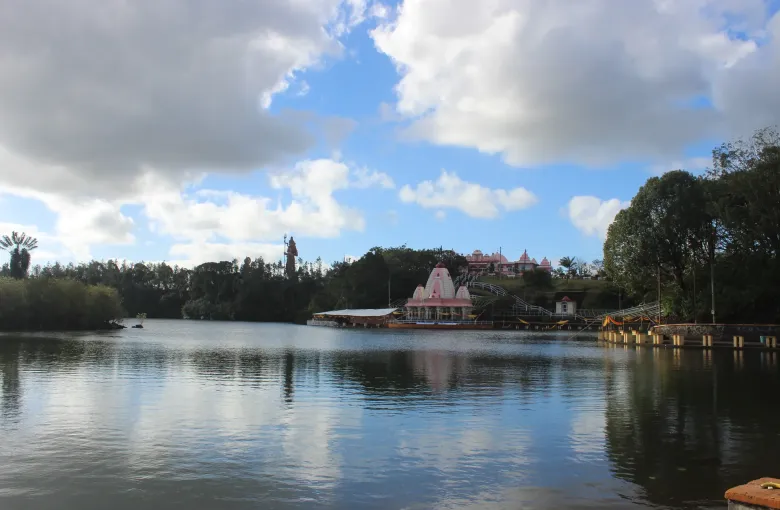 Temples and a tall standing statue reflected in the water of Ganga Talao, the sacred crater lake in the uplands of Mauritius.