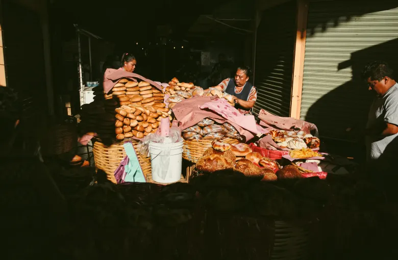 A bread stall piled with loaves and rolls inside the Mercado 20 de Noviembre in Oaxaca, with vendors behind the counter.