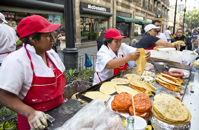 Cooks working a street taco stall in Mexico City, with tortillas on the griddle and fillings laid out along the counter.