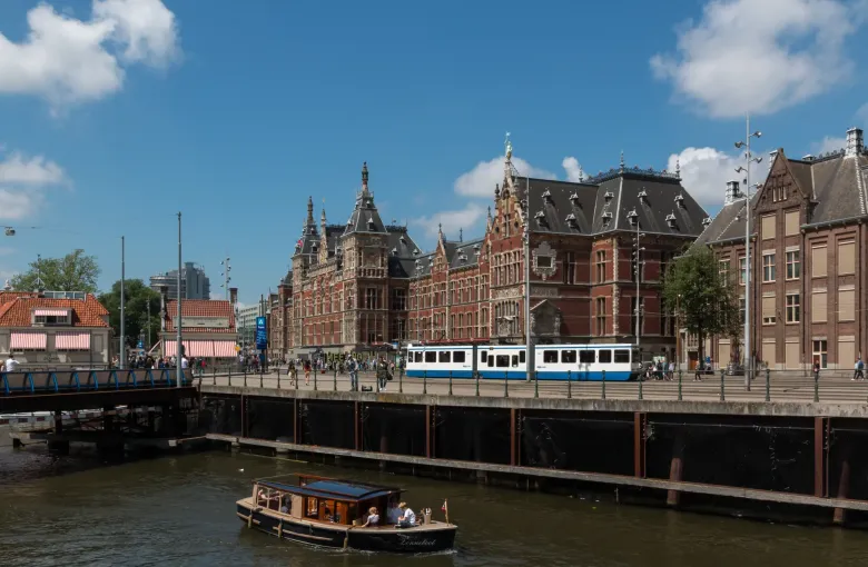A tram crossing in front of Amsterdam Centraal station while a canal boat passes below on the water.