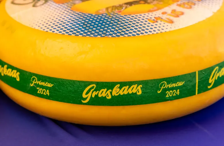 A yellow wax wheel of Dutch graskaas on a market stall, its green band printed in Dutch with the name and the year.