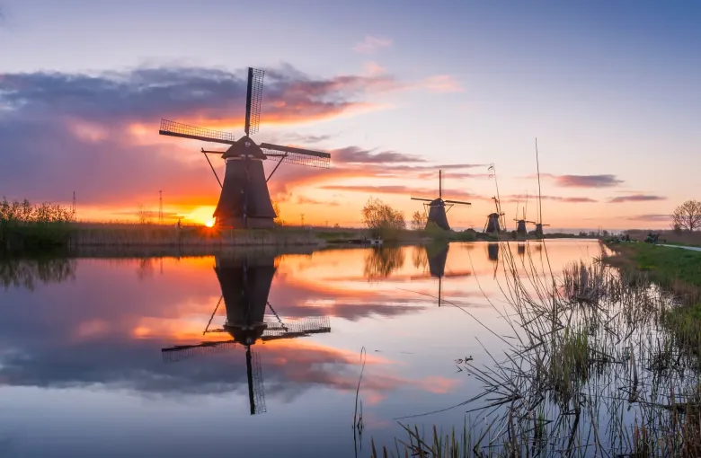 A line of drainage windmills at Kinderdijk receding into the distance along a dyke, mirrored in still water at sunrise.