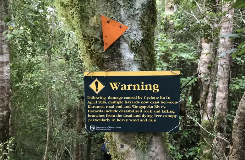 A Department of Conservation warning sign and an orange track marker fixed to a tree beside a bush track.