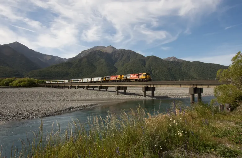 The TranzAlpine train crossing a long bridge over a braided river with mountains behind it.