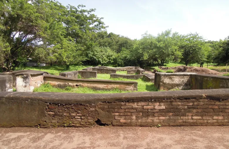 Low excavated foundation walls of Leon Viejo stand in cut grass among trees on level open ground.