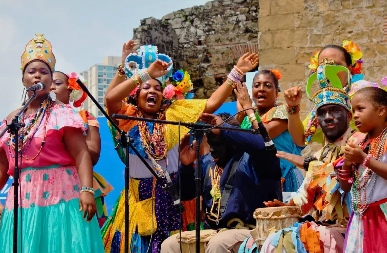 A Congo troupe singing and drumming on stage in bright layered dress before the stone ruins at Portobelo.