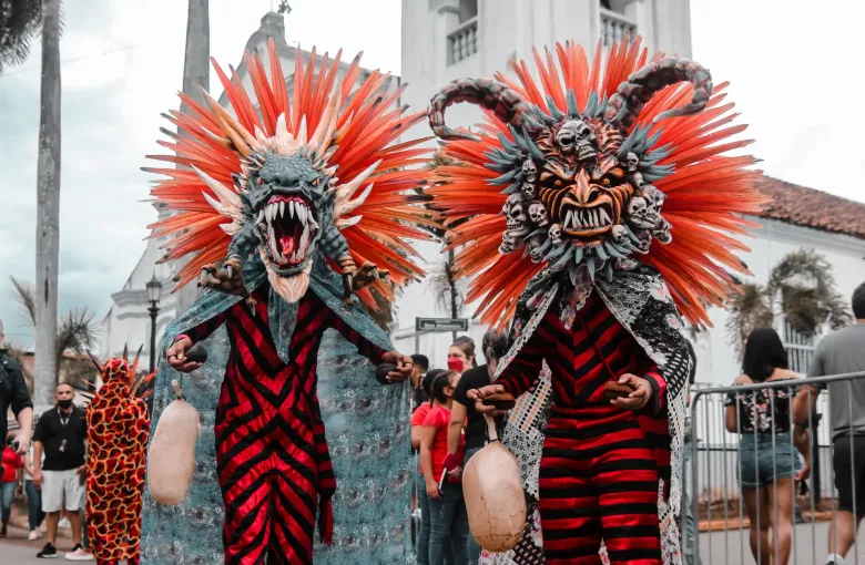 Two masked diablicos sucios dancers in spiked red headdresses and striped costume in a street by a church.