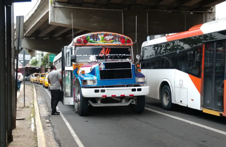 A hand painted diablo rojo bus beside a modern coach on a Panama City street under an overpass.