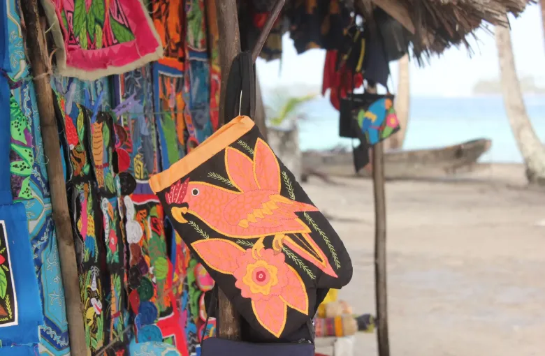 Hand stitched Guna molas hung for sale under a thatched shelter, with a dugout canoe behind.